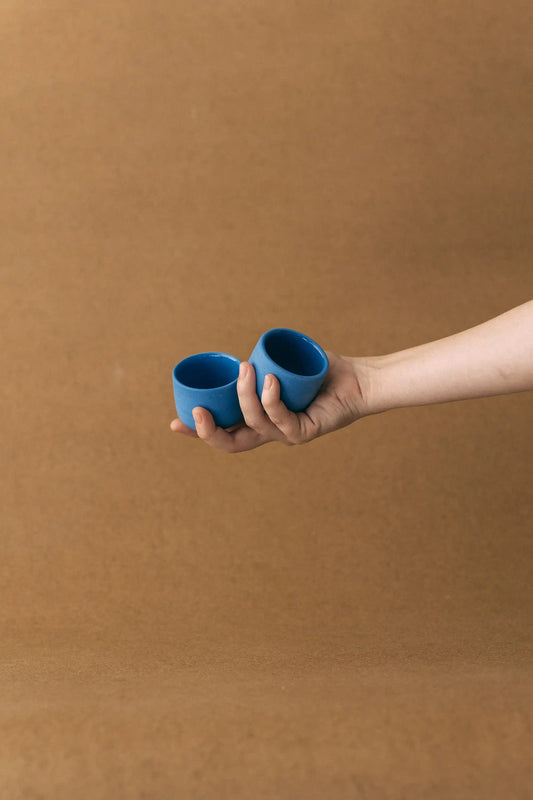 Ręka trzyma duet czarek ceramicznych PopUp 80ml w kolorze niebieskim na jasno brązowym tle