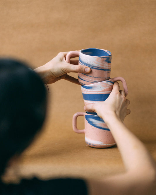 Trzy kubki ceramiczne z uchem Niebko, ustawione nierówno jeden na drugim, podtrzymywane przez dwie dłonie
