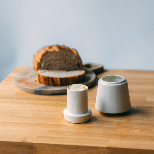 Drewniany blat, na którym stoi biała, nakrapiana maselniczka, której górna część wypełniona jest masłem. W tyle chleb na desce do krojenia.