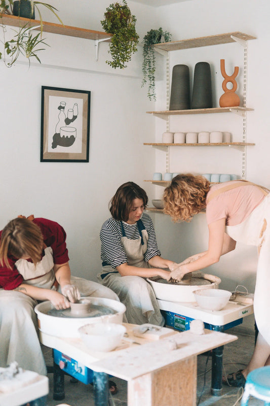 Dwie uczestniczki, instruowane przez ceramiczkę prowadząca warsztaty, pracujące przy kołach garncarskich w przytulnej pracowni z roślinami i półkami ceramiki - miesięczny kurs garncarski