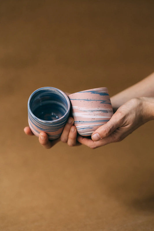 Dłonie prezentujące dwie ceramiczne czarki w marmurkowym kolorze niebieskim, białym i różowym