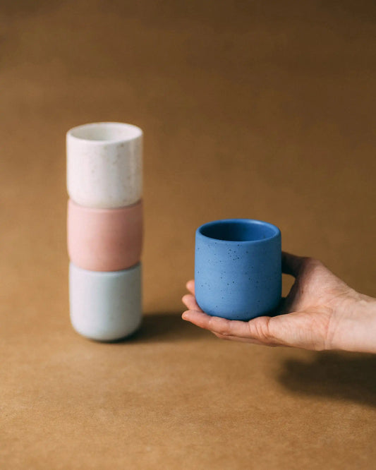 Ręka trzymająca niebieski, nakrapiany ceramiczny kubek obok wieży ustawionej z tych samych kubków w kolorze białym, różowym i szałwiowym