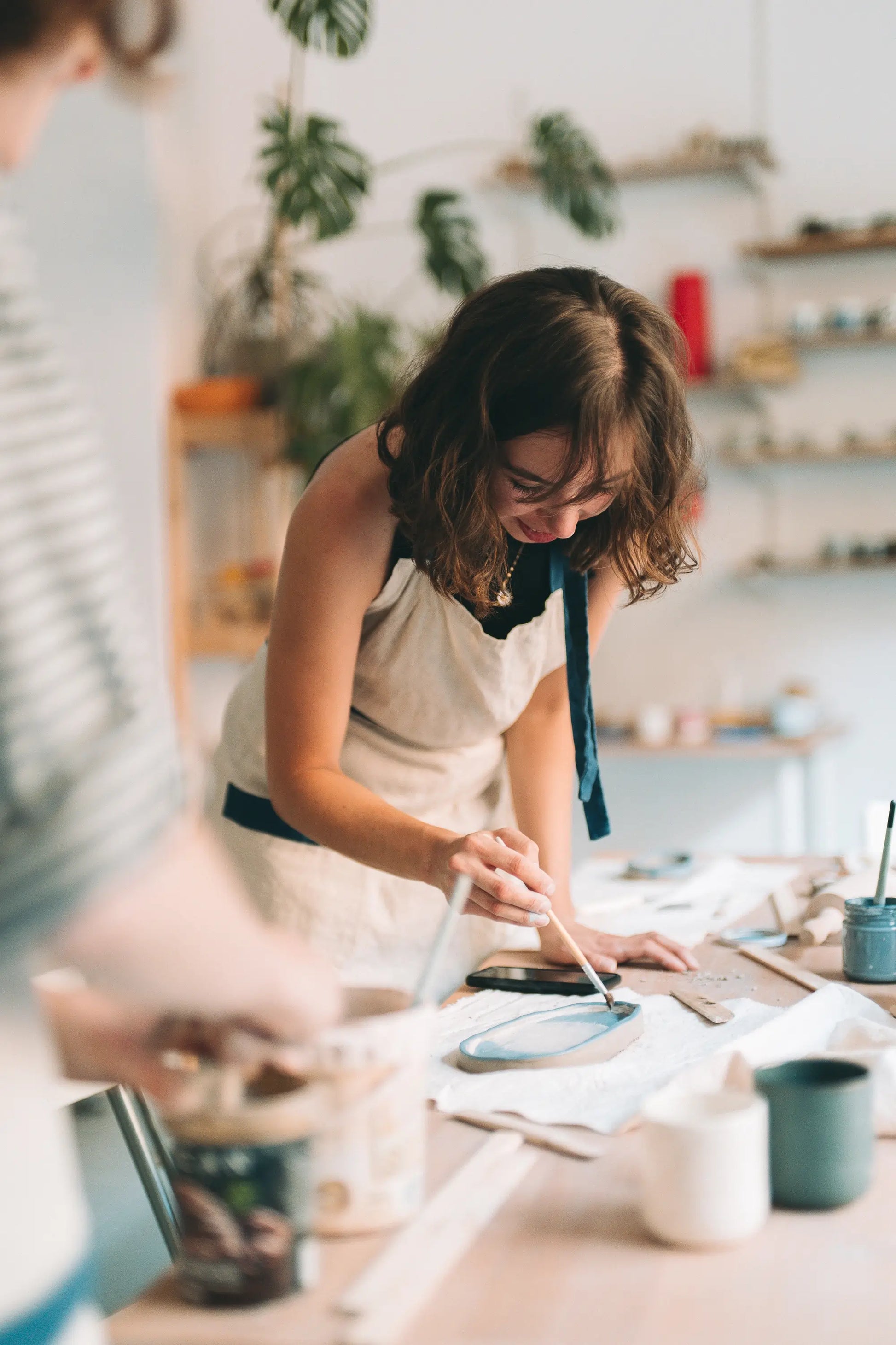 Uczestniczka warsztatu w fartuchu malująca talerz pędzlem w jasnej pracowni z roślinami - voucher skrócony kurs lepienia 2 spotkania