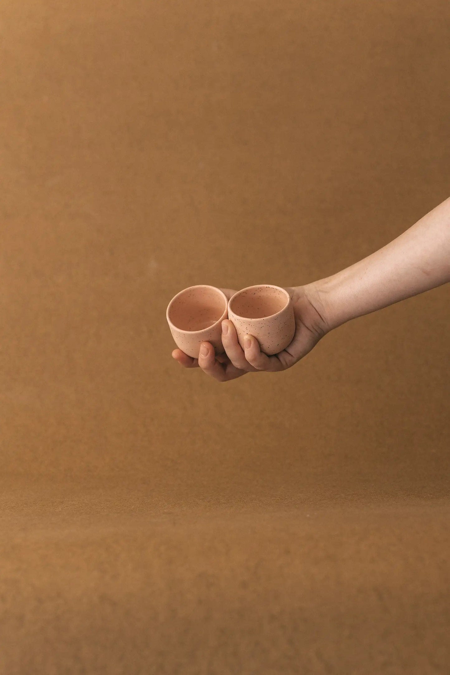 Ręka trzyma duet czarek ceramicznych PopUp 80ml w kolorze różowym na jasno brązowym tle