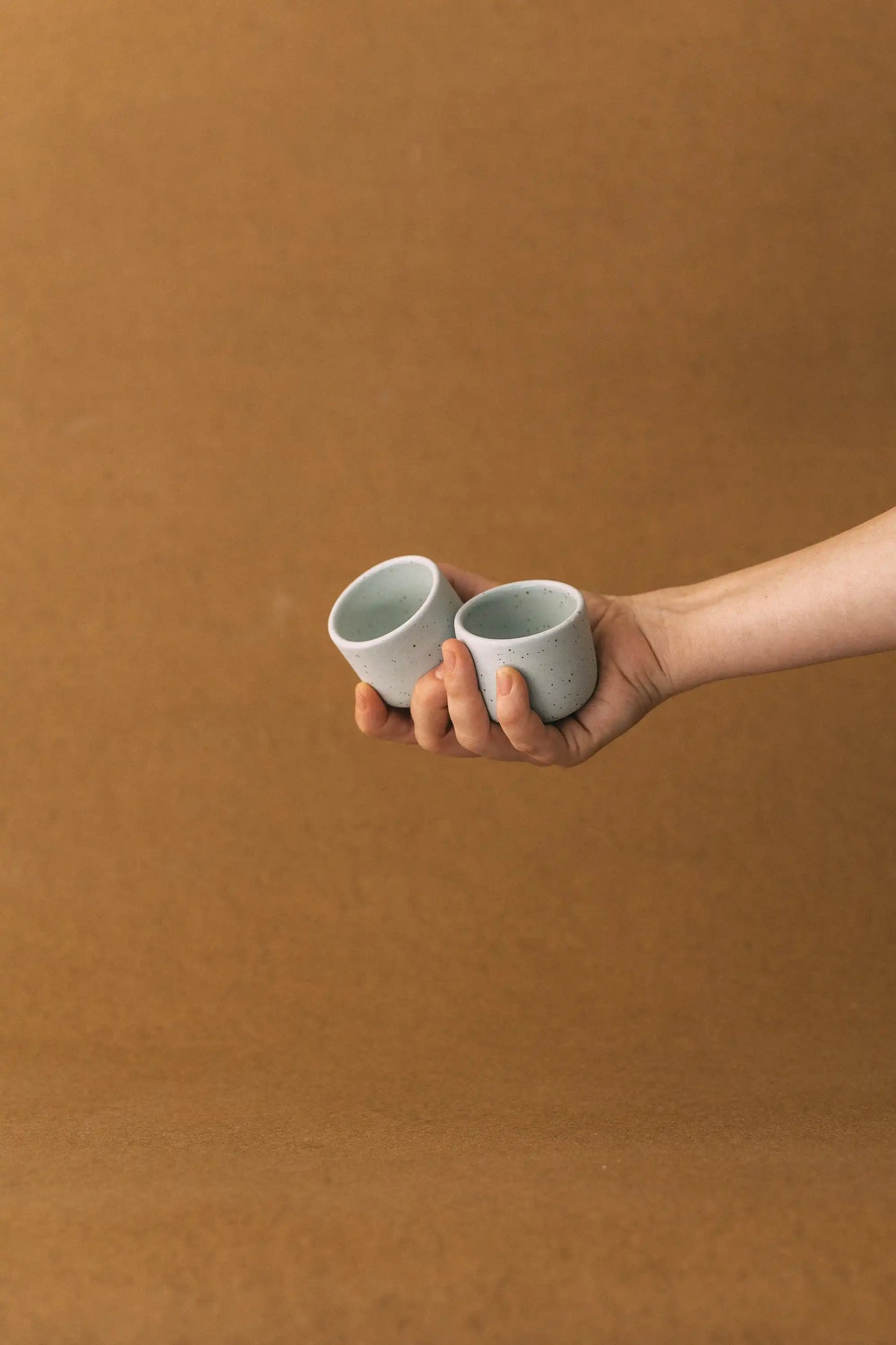 Ręka trzyma duet czarek ceramicznych PopUp 80ml w kolorze szałwiowym na jasno brązowym tle