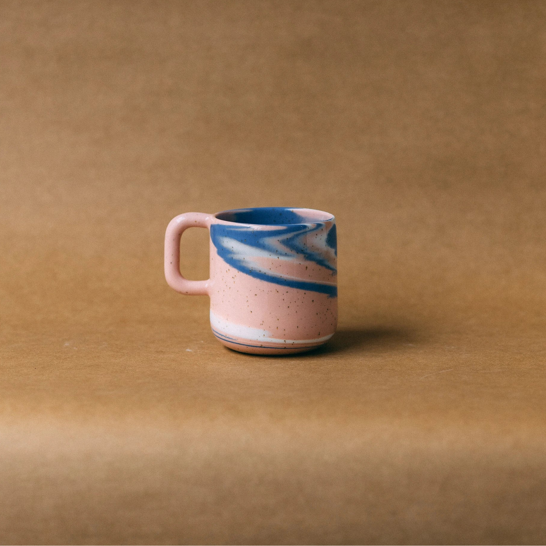 Kubek ceramiczny z uchem Niebko, różowo-niebiesko-biały, ustawiony na jasno brązowym tle - widok z boku