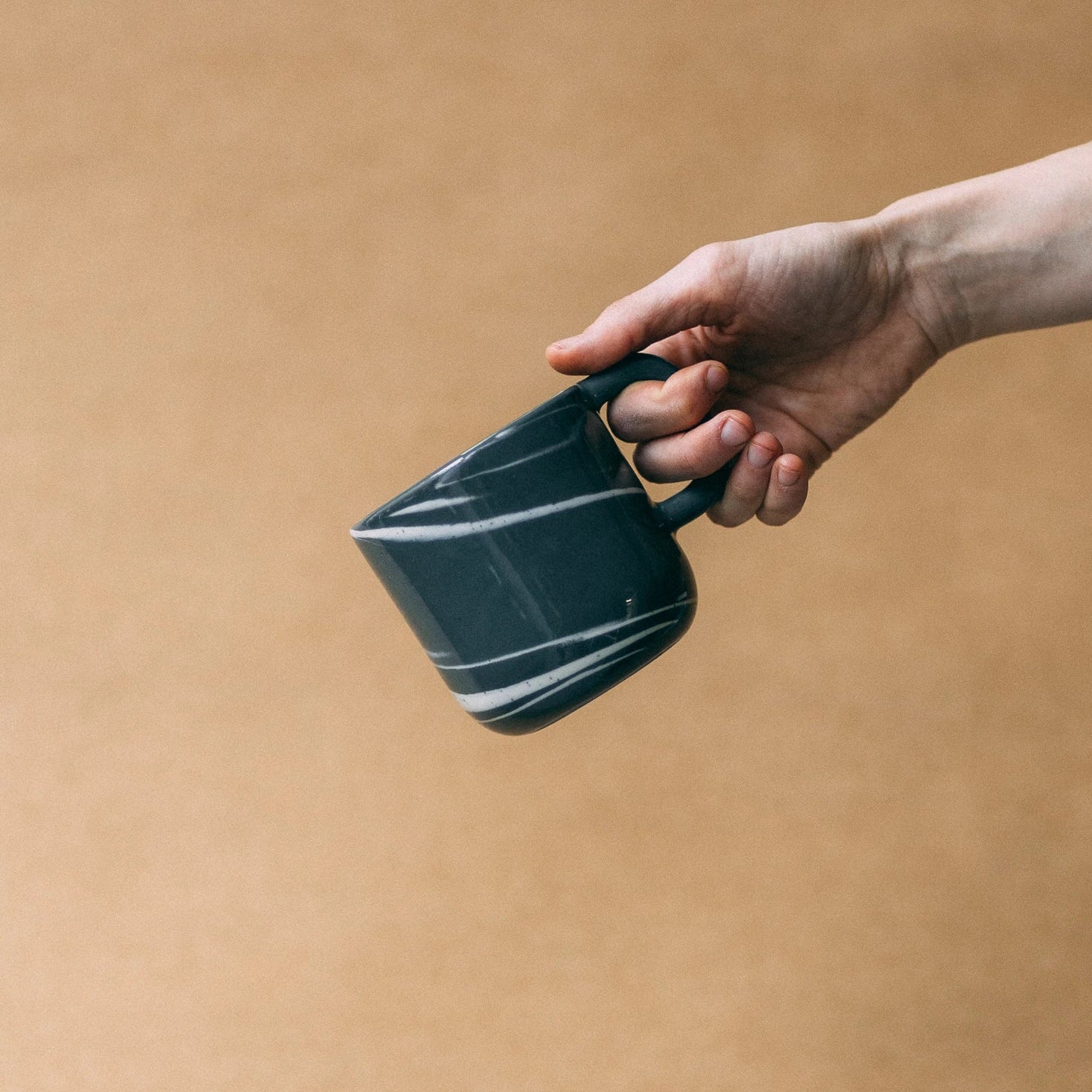 Kubek ceramiczny z uchem Smog grafitowy z elementami bieli, trzymany w powietrzu przez dłoń na jasno brązowym tle - widok z boku