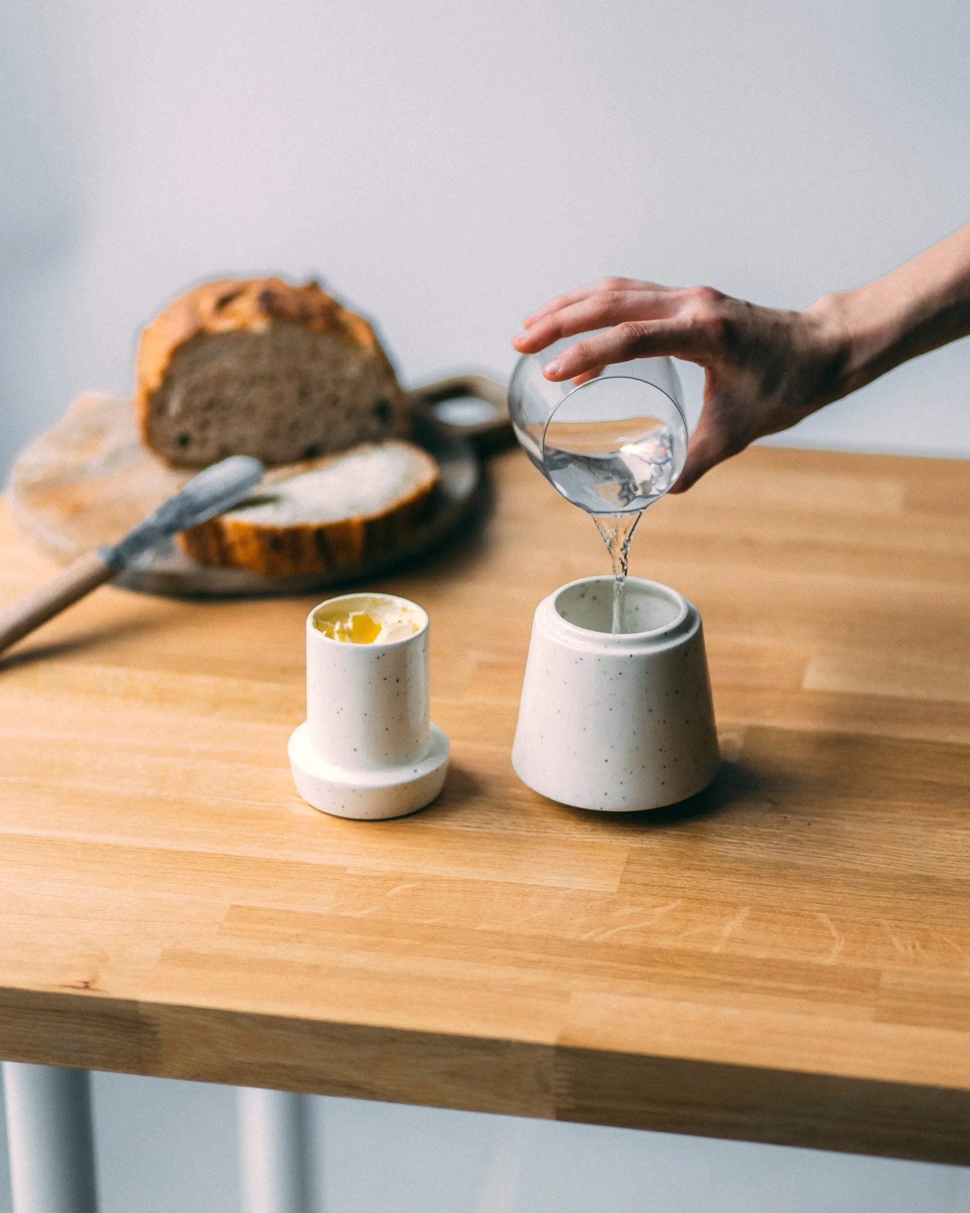 Drewniany blat, na którym stoi biała, nakrapiana maselniczka, jej górna część wypełniona jest masłem, a do dolnej ktoś nalewa wody ze szklanki. W tyle chleb na desce do krojenia i metalowy nóż do masła.
