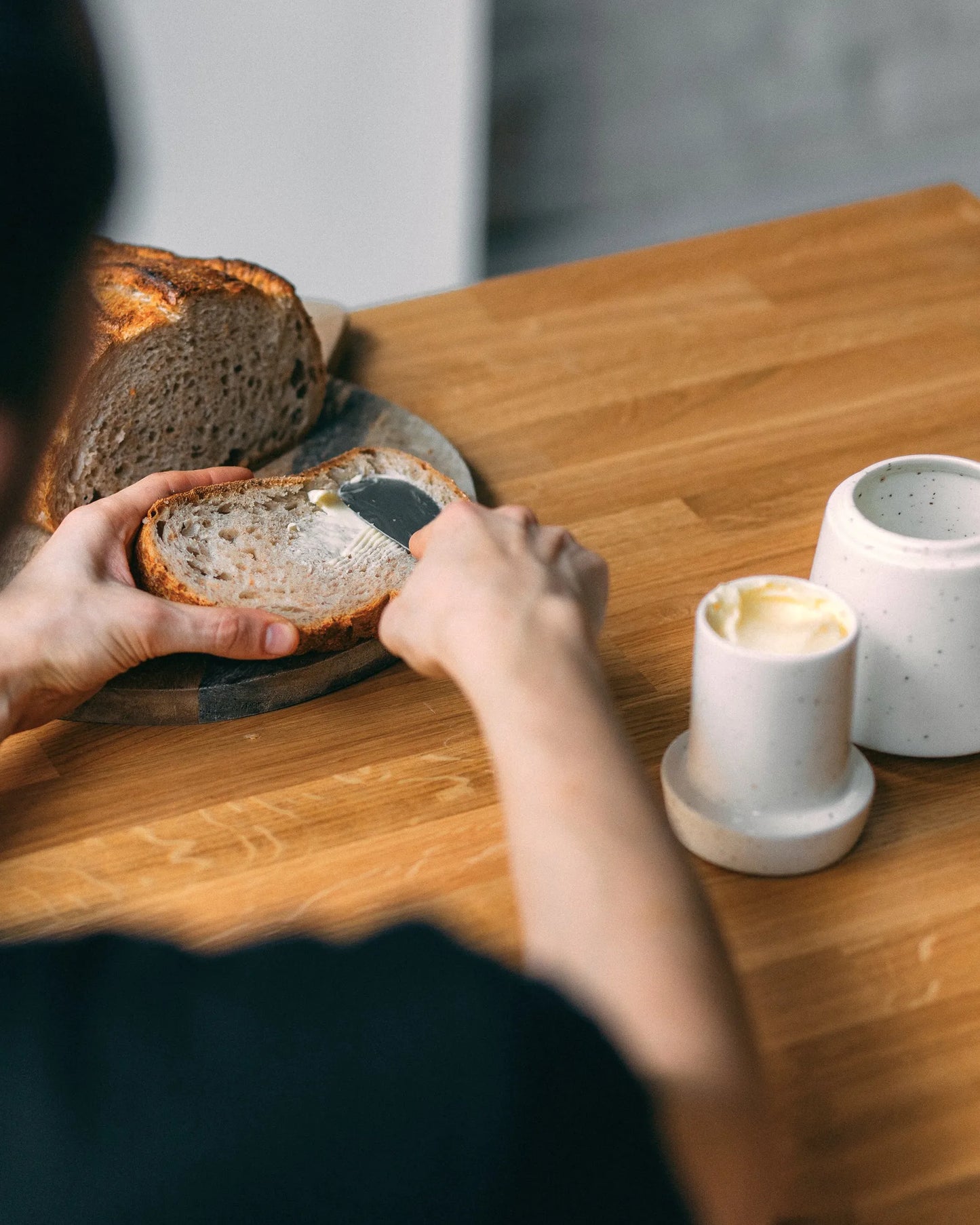 Smarowanie masłem kromki chleba, po prawej stronie widoczna maselniczka z górną częścią wypełnioną wodą.