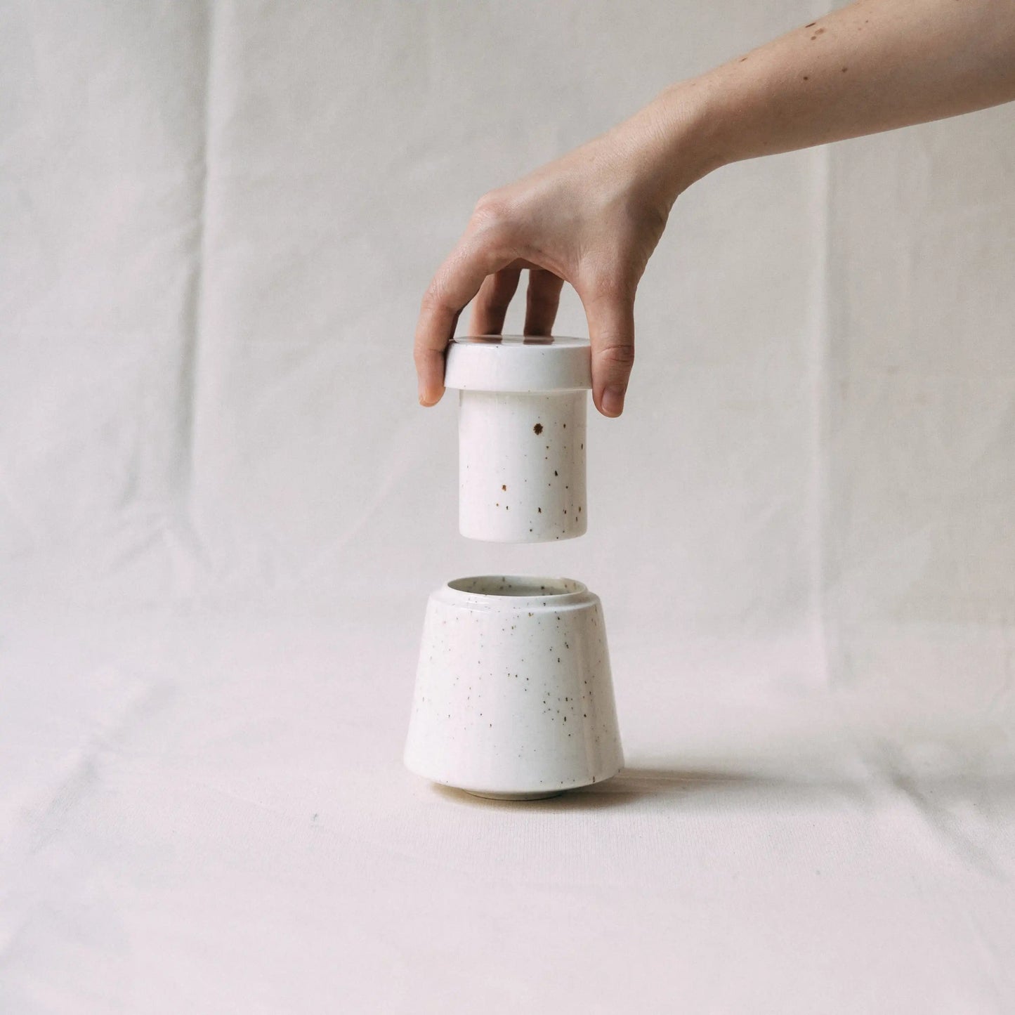 Dłoń unosząca górną część maselniczki wodnej ceramicznej MUFKA w kolorze białym
