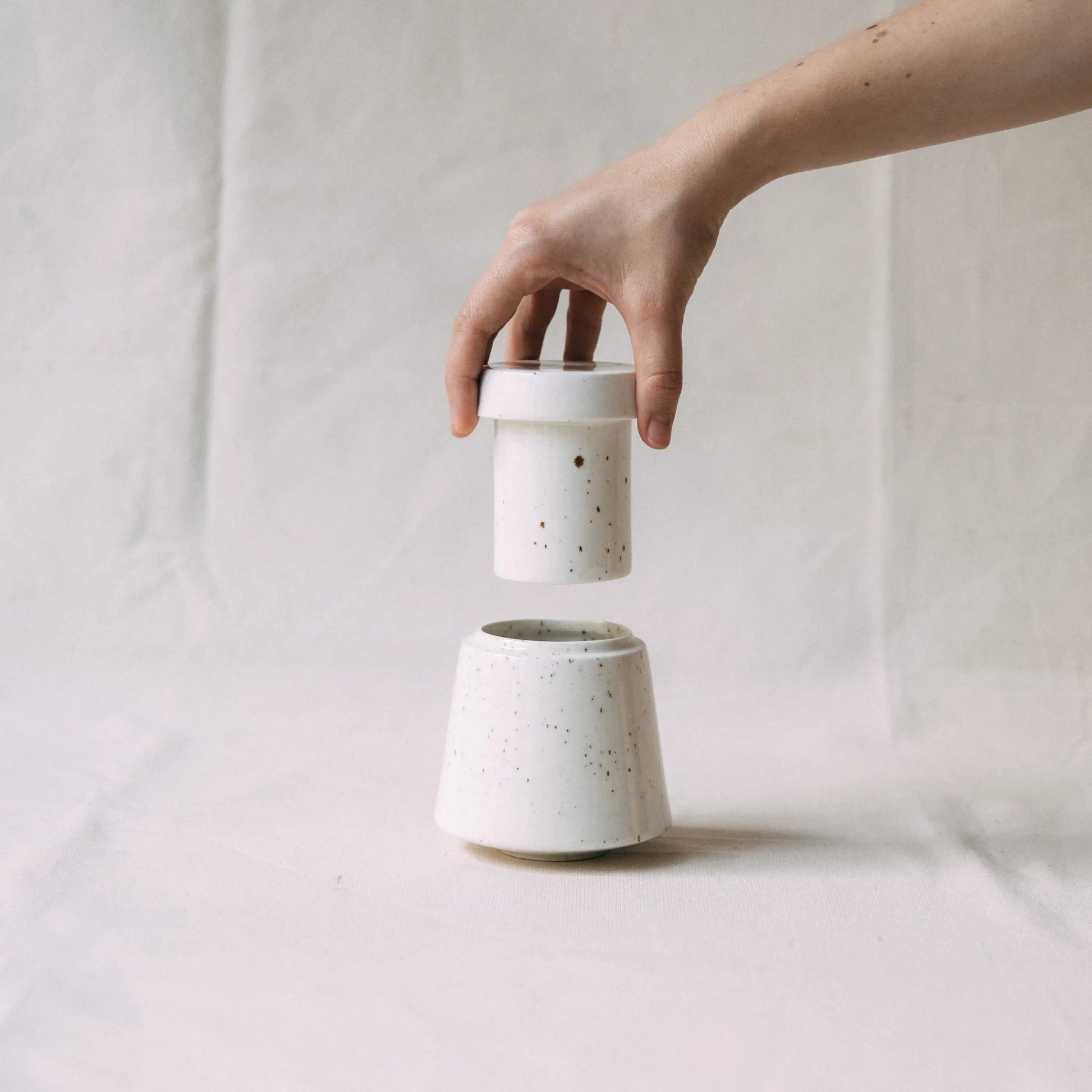 Dłoń unosząca górną część maselniczki wodnej ceramicznej MUFKA w kolorze białym