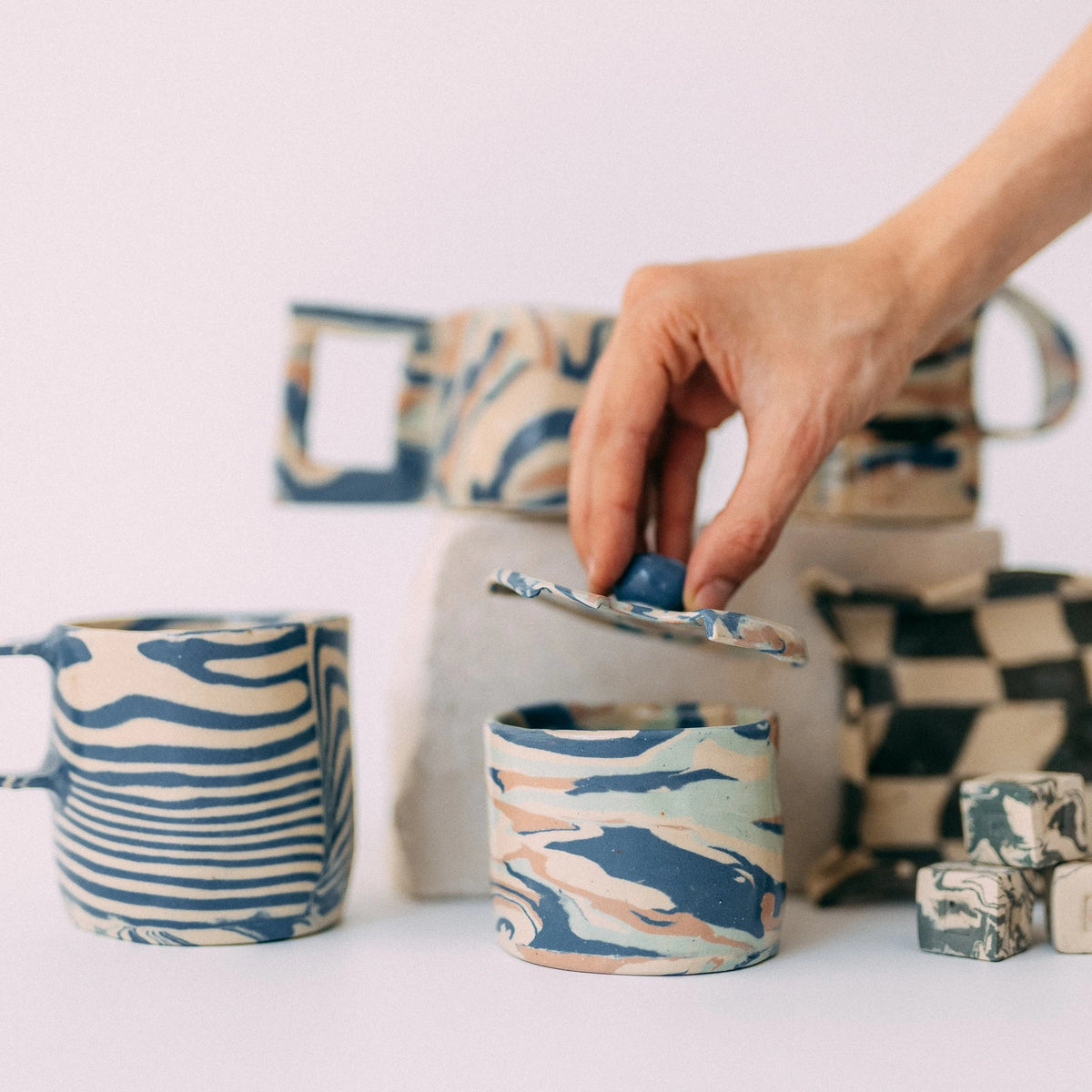 Kilka ceramicznych prac warsztatowych. Na pierwszym planie małe ceramiczne pudełko z pokrywką, którą podnosi dłoń.