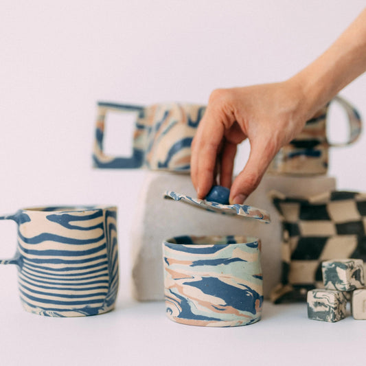 Kilka ceramicznych prac warsztatowych. Na pierwszym planie małe ceramiczne pudełko z pokrywką, którą podnosi dłoń.