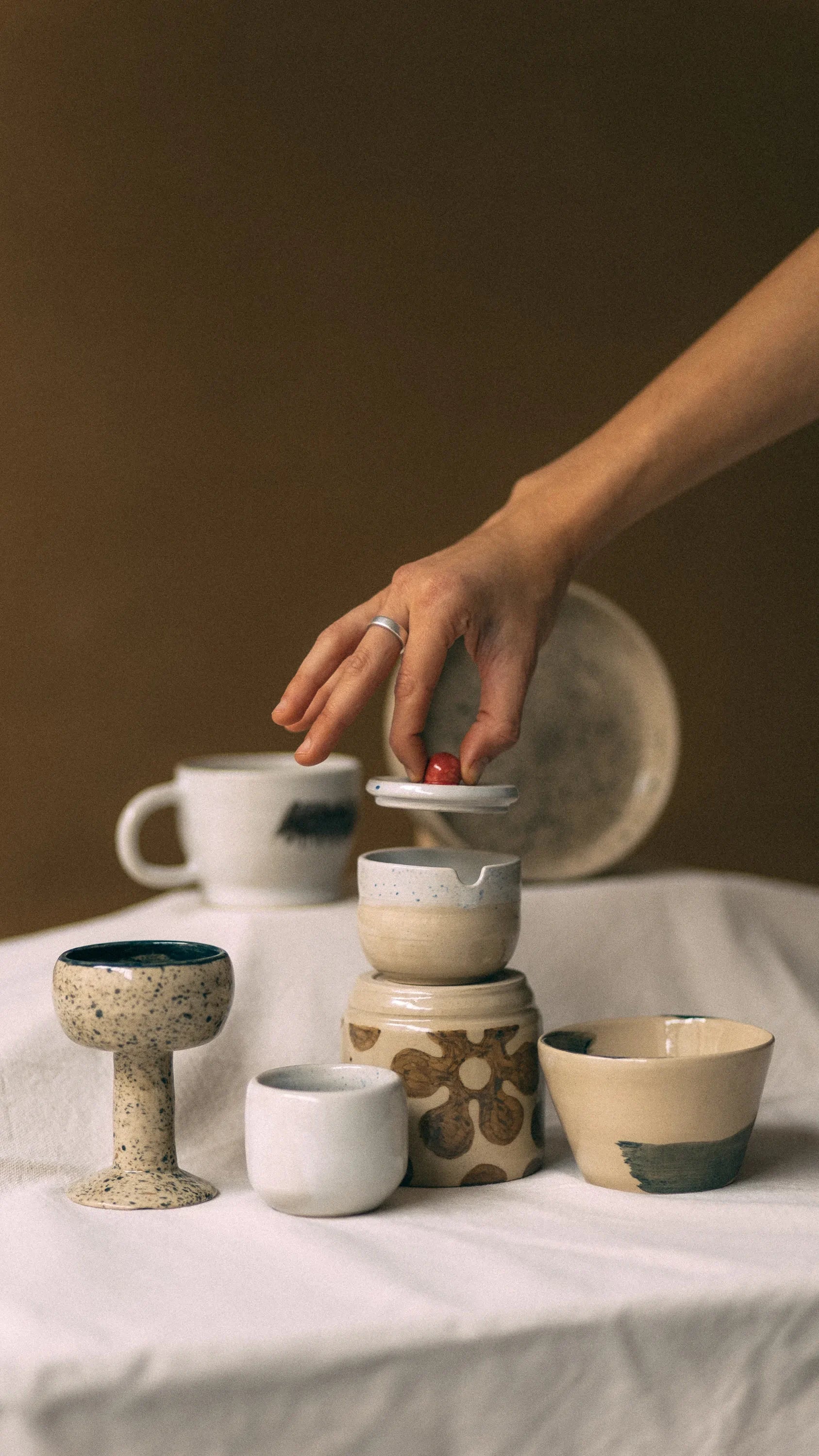 Dłoń trzymając za czerwony uchwyt podnosi ceramiczną pokrywkę od cukierniczki otoczonej innymi ceramicznymi przedmiotami codziennego użytku  - miesięczny kurs garncarski