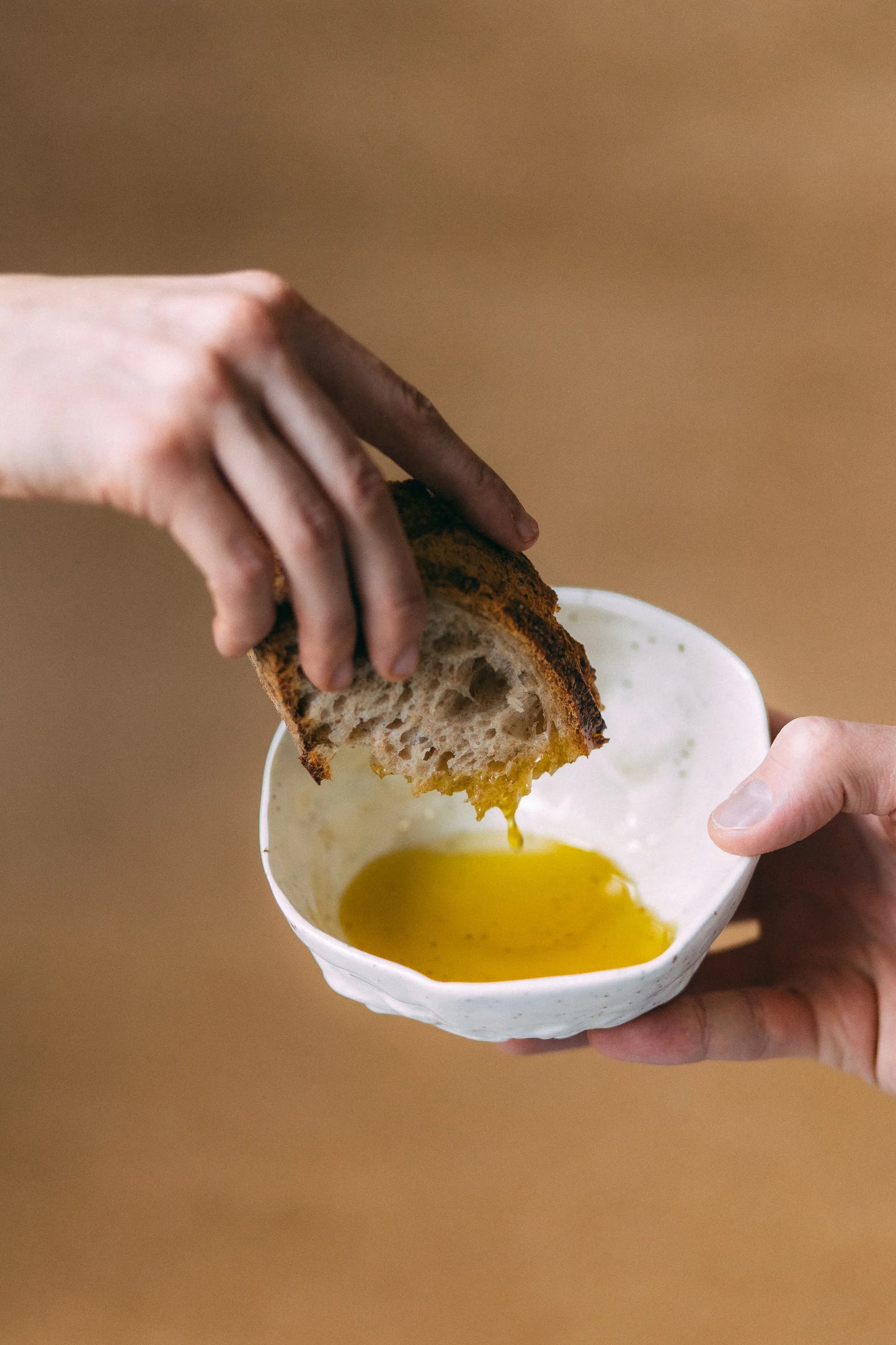 Jedna dłoń podaje białą miskę wypełnioną małą ilością oliwy, w której z drugiej strony inna ręka macza kromkę chleba.