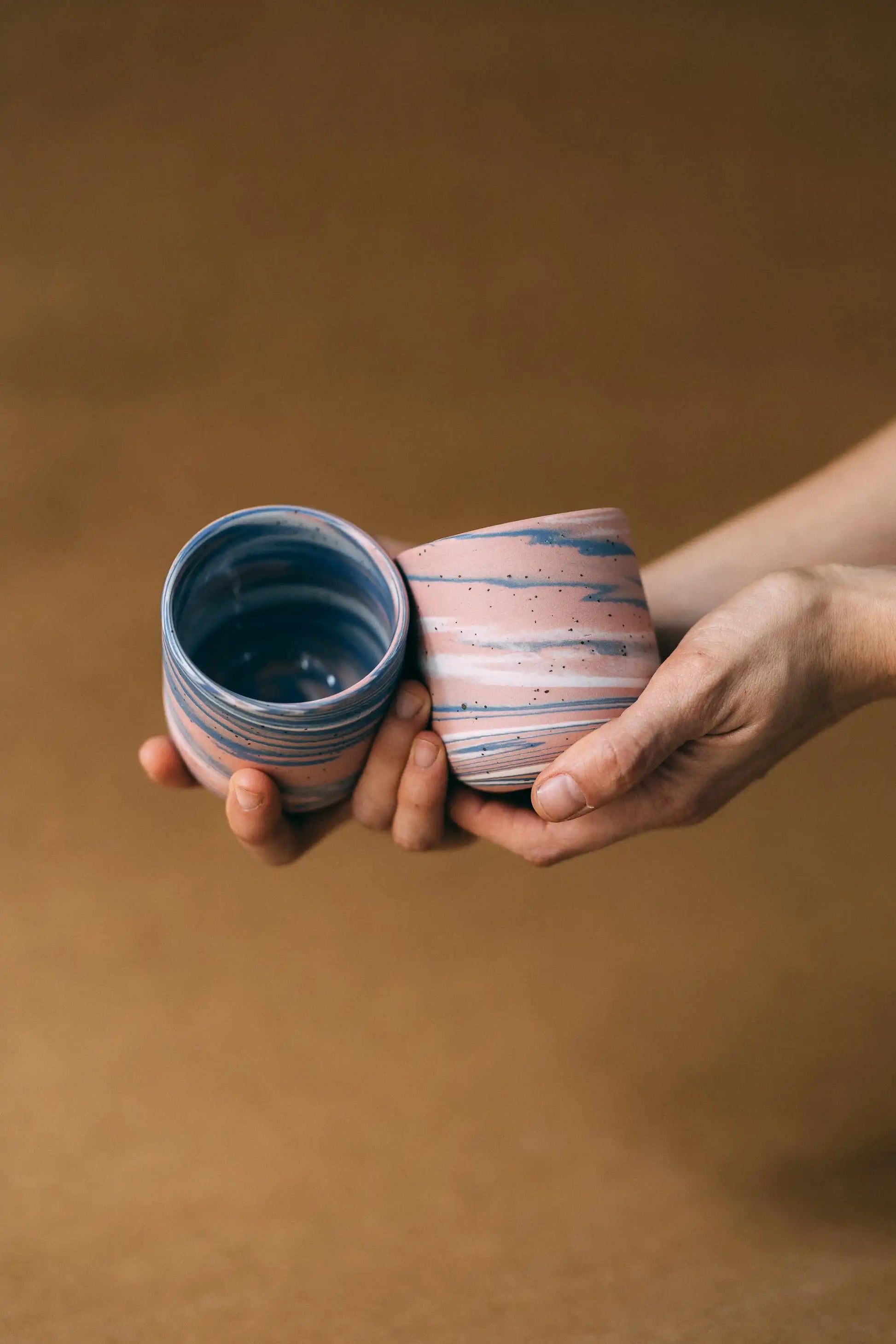 Dłonie prezentujące dwie ceramiczne czarki w marmurkowym kolorze niebieskim, białym i różowym