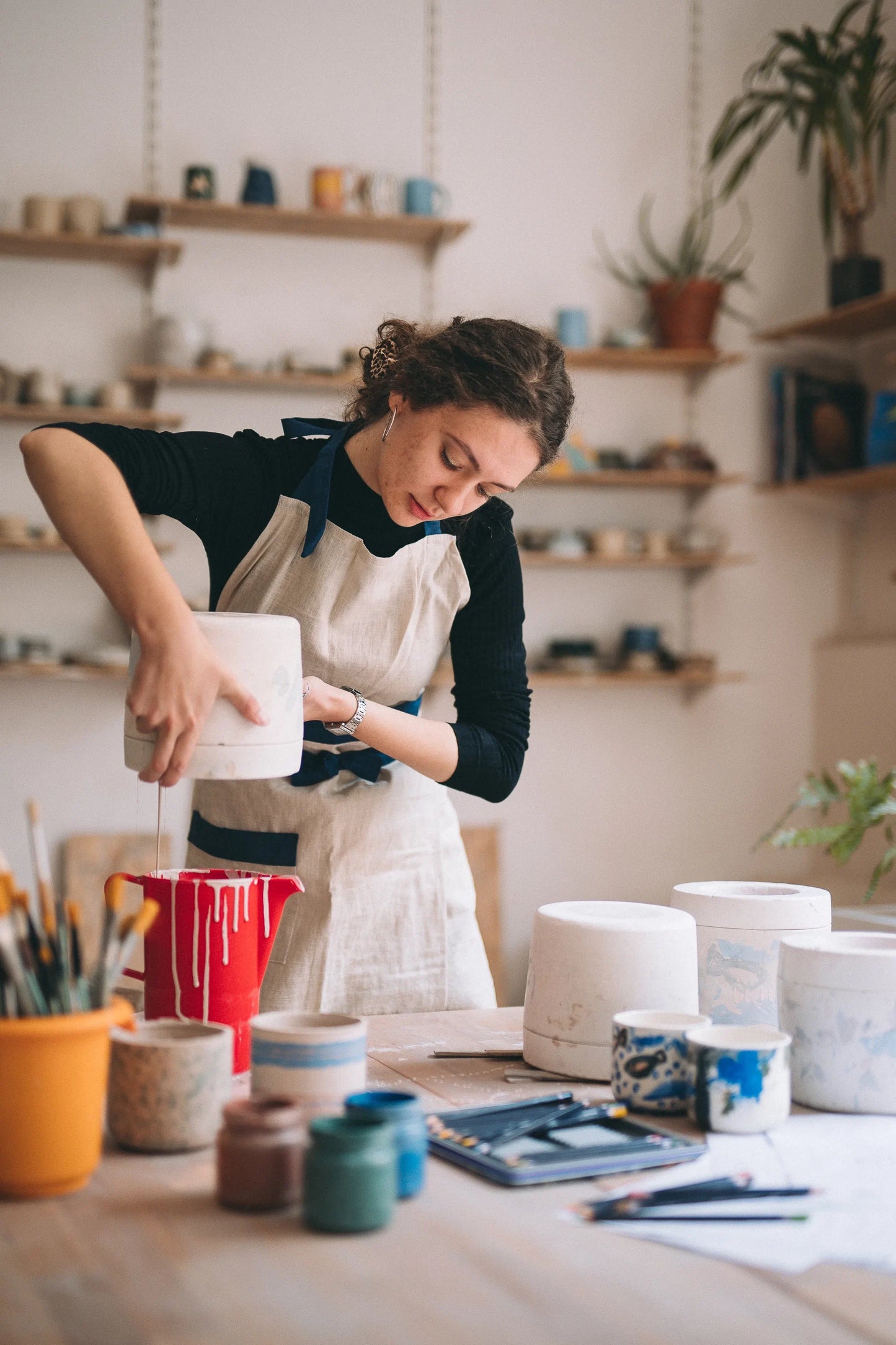 Ceramiczka odlewająca płynną porcelanę z pojemnika do formy gipsowej otoczona kolorowymi kubkami - warsztat odlewania porcelany