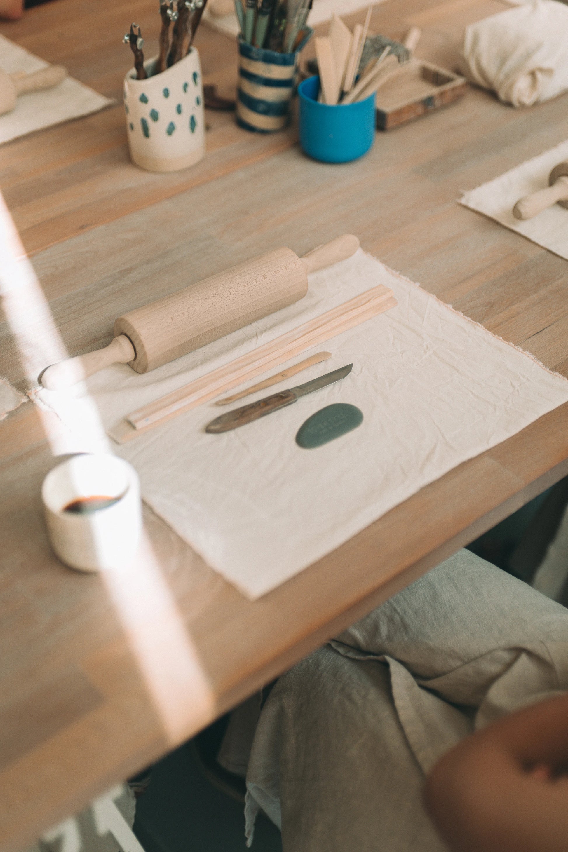 Fragment stołu, na którym znajdują się przedmioty przygotowane do warsztatu lepienia z gliny, takie jak ściereczka, drewniany wałek i narzędzia, nożyk oraz gumowa cyklina.