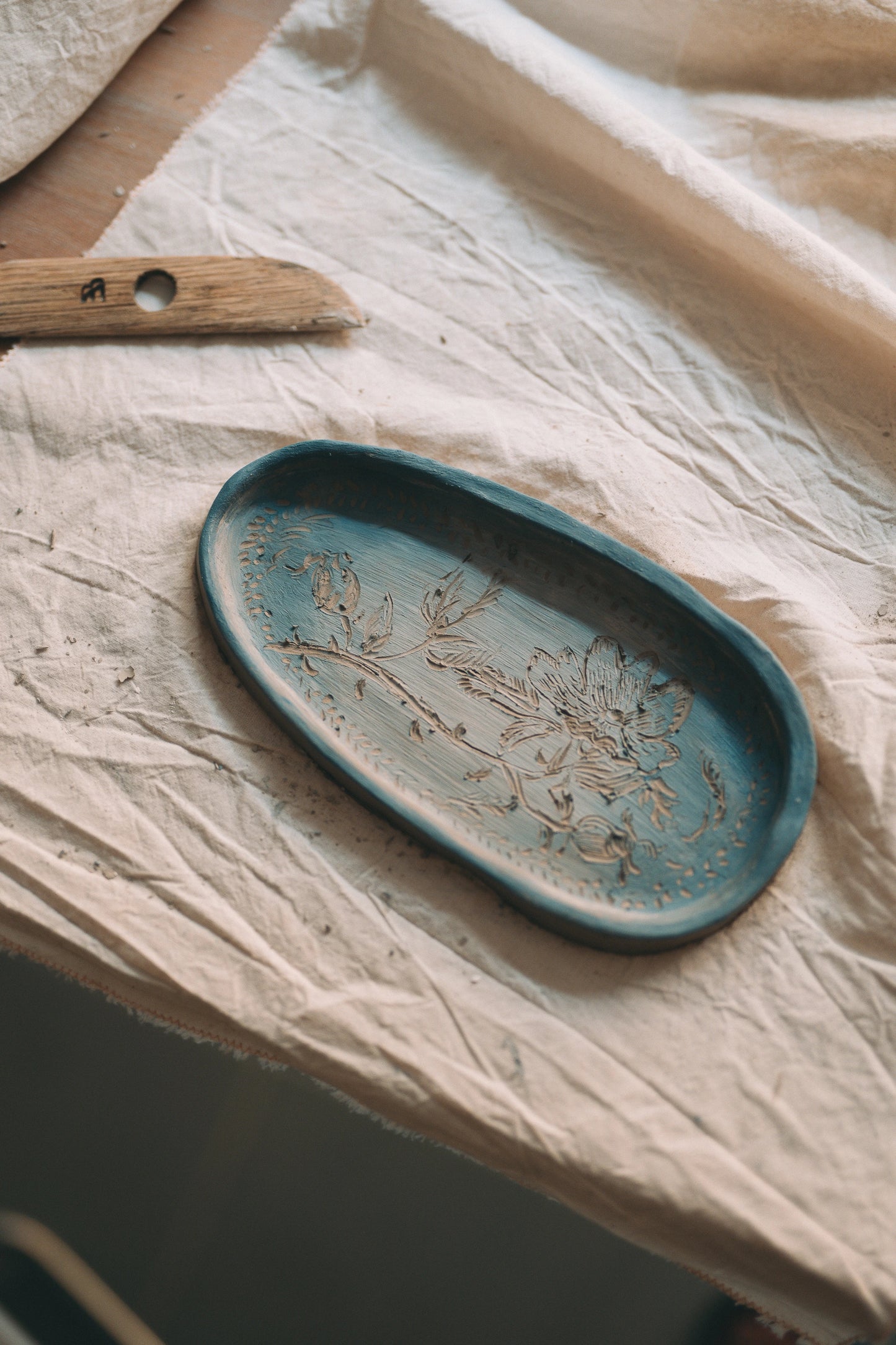 Patera ulepiona z gliny z płaskorzeźbą z motywem roślinnym, pomalowana na niebiesko, a obok drewniane narzędzie do lepienia