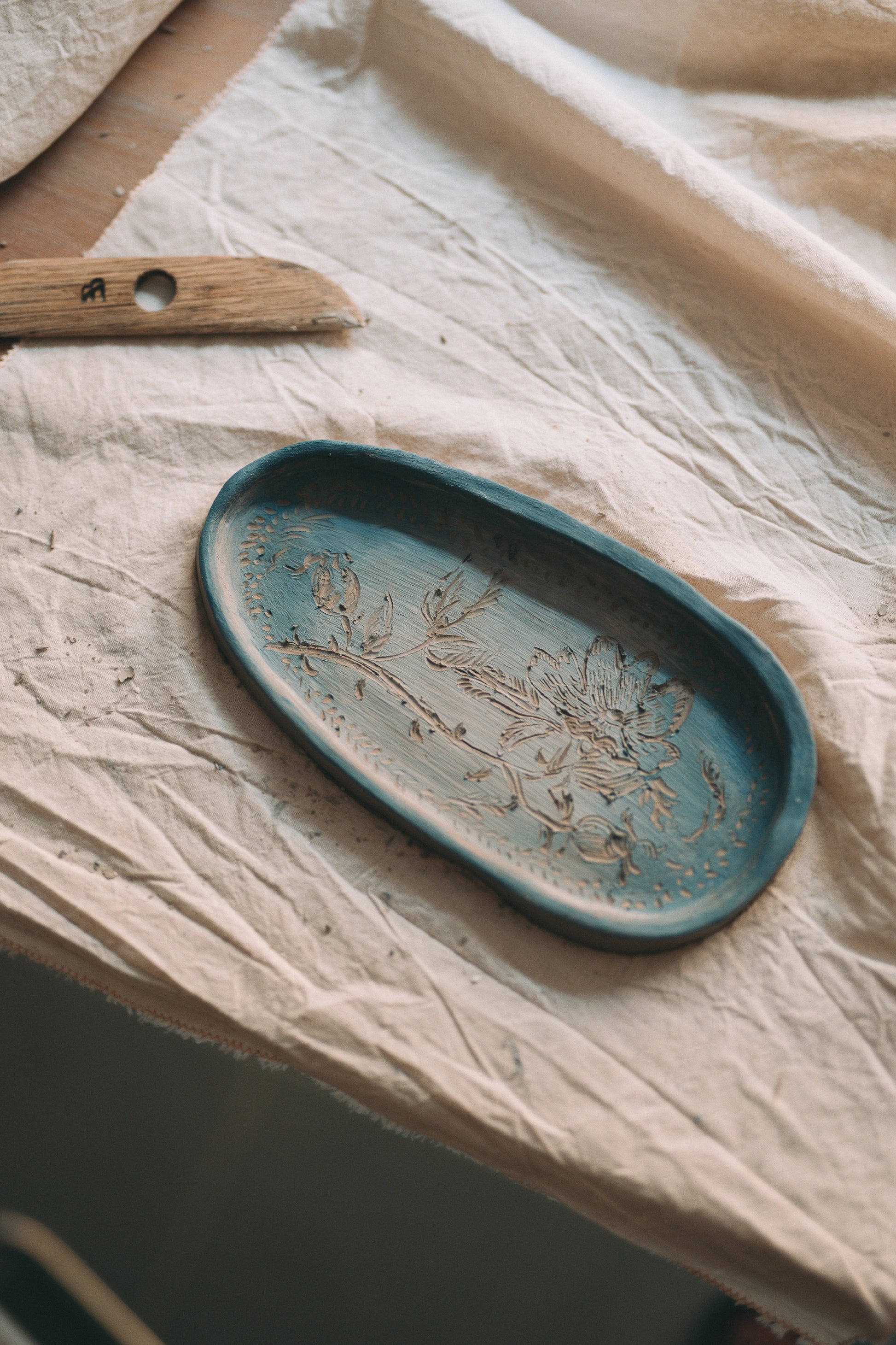 Patera ulepiona z gliny z płaskorzeźbą z motywem roślinnym, pomalowana na niebiesko, a obok drewniane narzędzie do lepienia