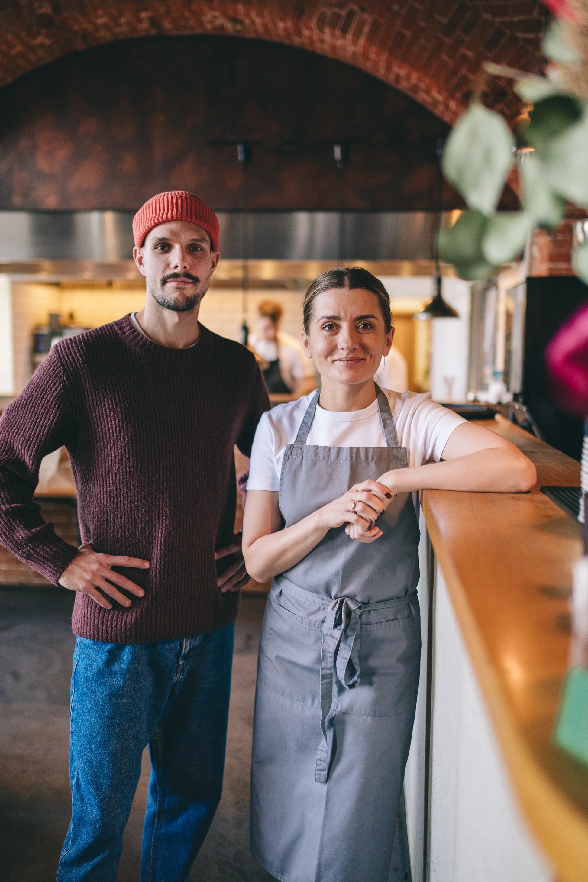 Uśmiechnięta kobieta wsparta łokciem o ladę i mężczyzna  z rękoma opartymi na biodrach stojący na tle restauracji z ceglastym sufitem.