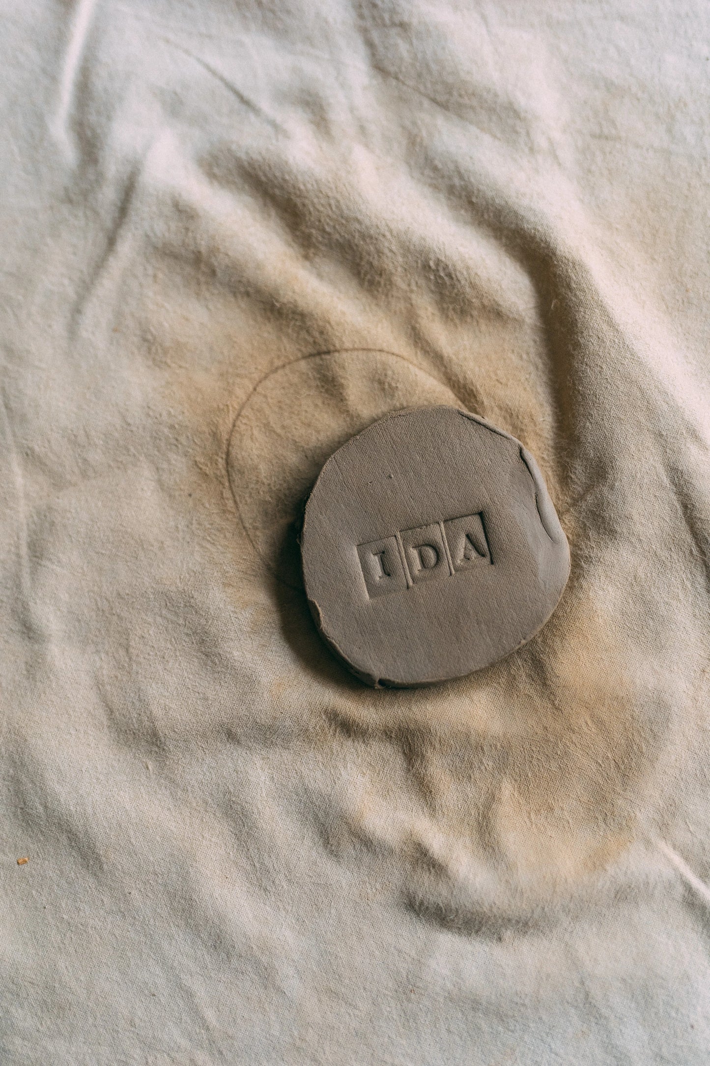 Płaskie kółko surowej gliny, w którym odbite jest słowo IDA, leżące na beżowej ściereczce.