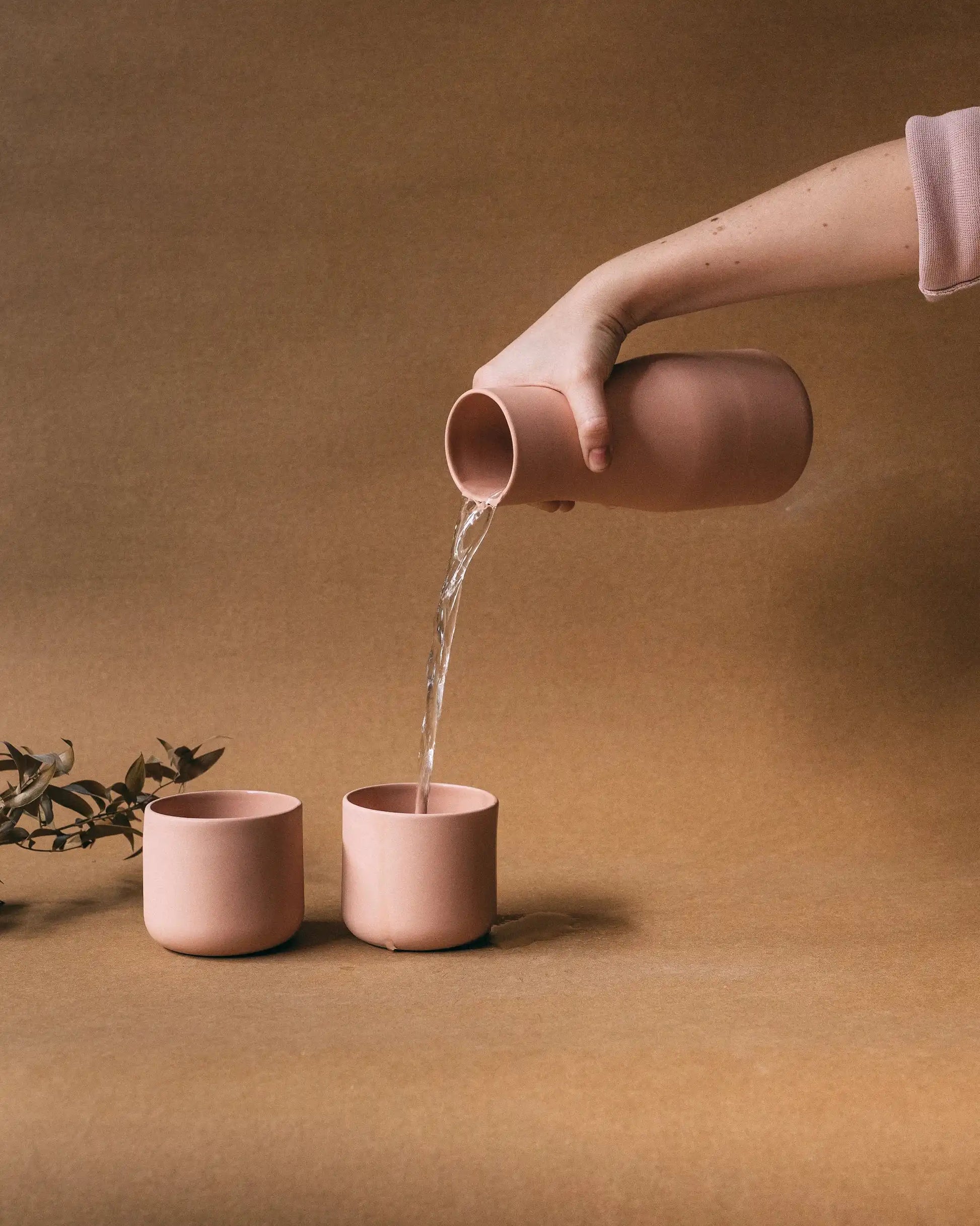 Ręka nalewająca wodę z karafki do jednej z dwóch filiżanek. Karafka i filiżanki w matowym pudrowym różowym kolorze - kolekcja ZakwasTAP