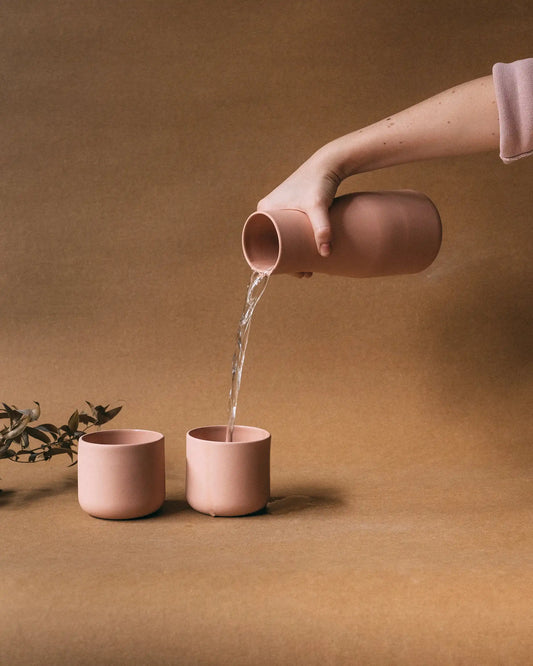 Ręka nalewająca wodę z karafki do jednej z dwóch filiżanek. Karafka i filiżanki w matowym pudrowym różowym kolorze - kolekcja ZakwasTAP